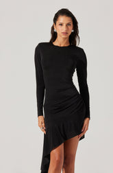 A woman is wearing a long-sleeved, fitted black dress with a round neckline. The dress features a textured fabric and an asymmetrical ruffle detail that runs along the hem, creating a high-low effect. She has dark, wavy hair and is accessorized with square earrings.